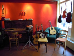 Gitarrenunterricht Wuppertal, Gitarrenzentrum. Photo vom Café Esperanza. rote Wand mit einem Charango. Davor ein altes Klavier, ein grüner Sessel, auf dem eine Gitarre liegt. 2 weitere Gitarren stehen im Gitarrenständer. 3 Stühle im Vordergrund, einer am Klavier, zwei zum Photographen ausgerichtet. Dazwischen Bongos. Im Schaufenster rechts sieht man 3 Gitarren hängen.